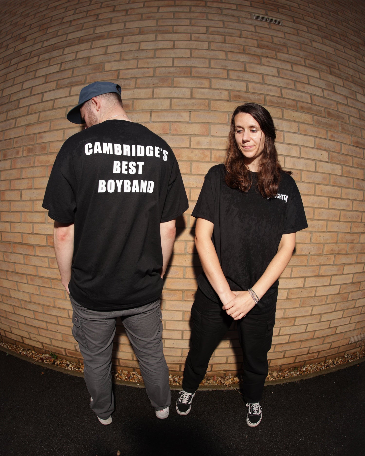 Two people wearing black BANGKITAN  t-shirts with text, standing against a brick wall.