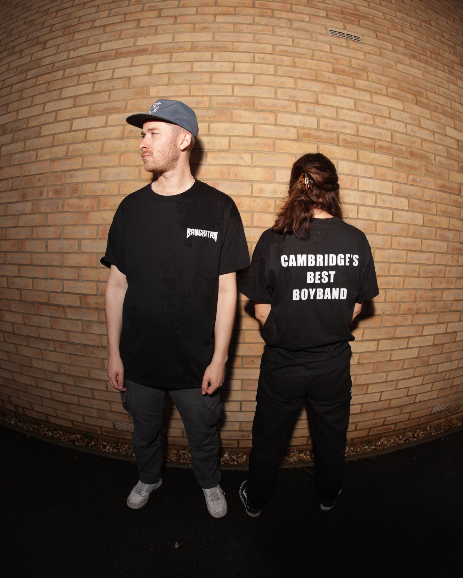 Two people wearing black BANGKITAN t-shirts standing in front of a brick wall