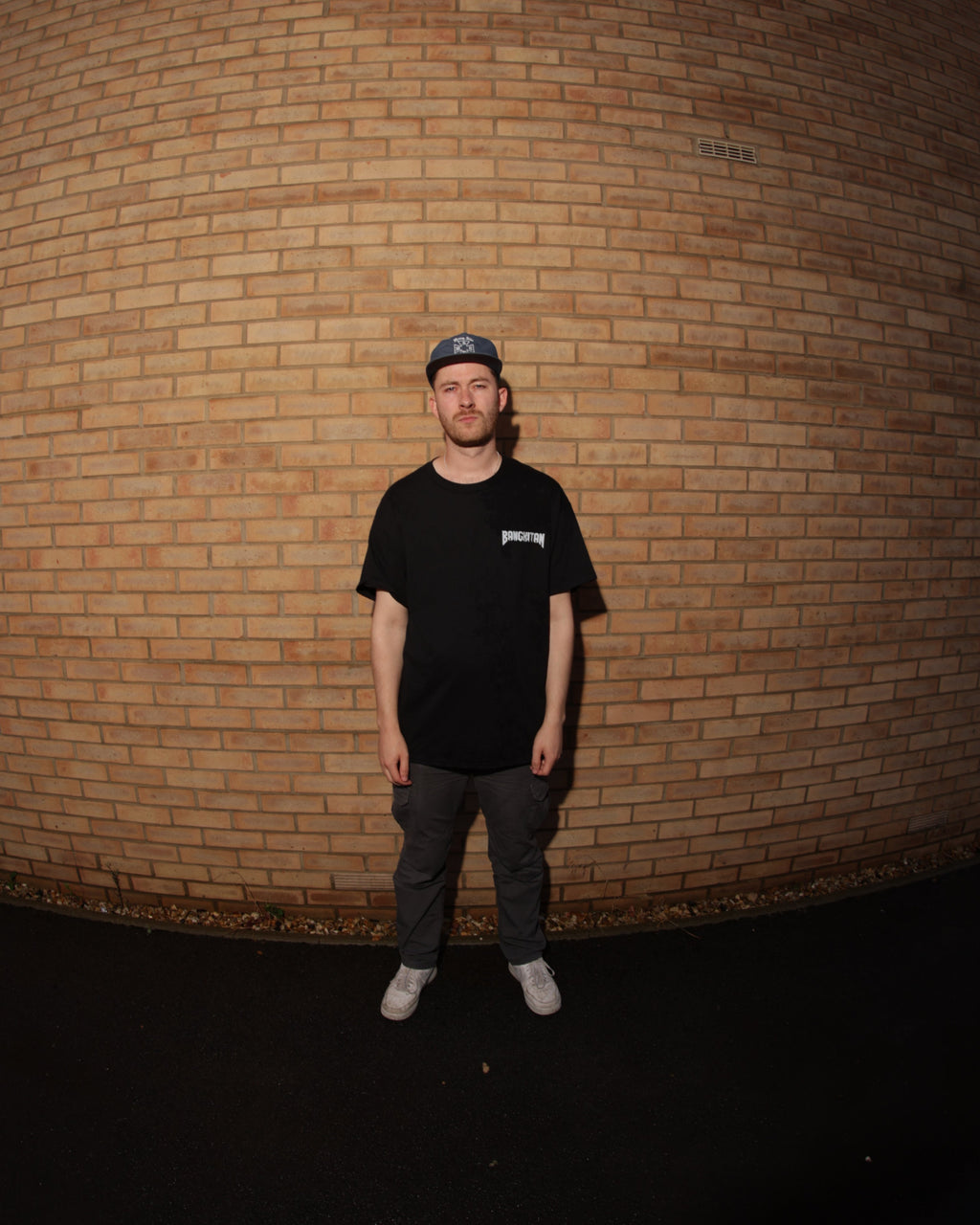 Man wearing a black BANGKITAN t-shirt and cap standing against a brick wall.