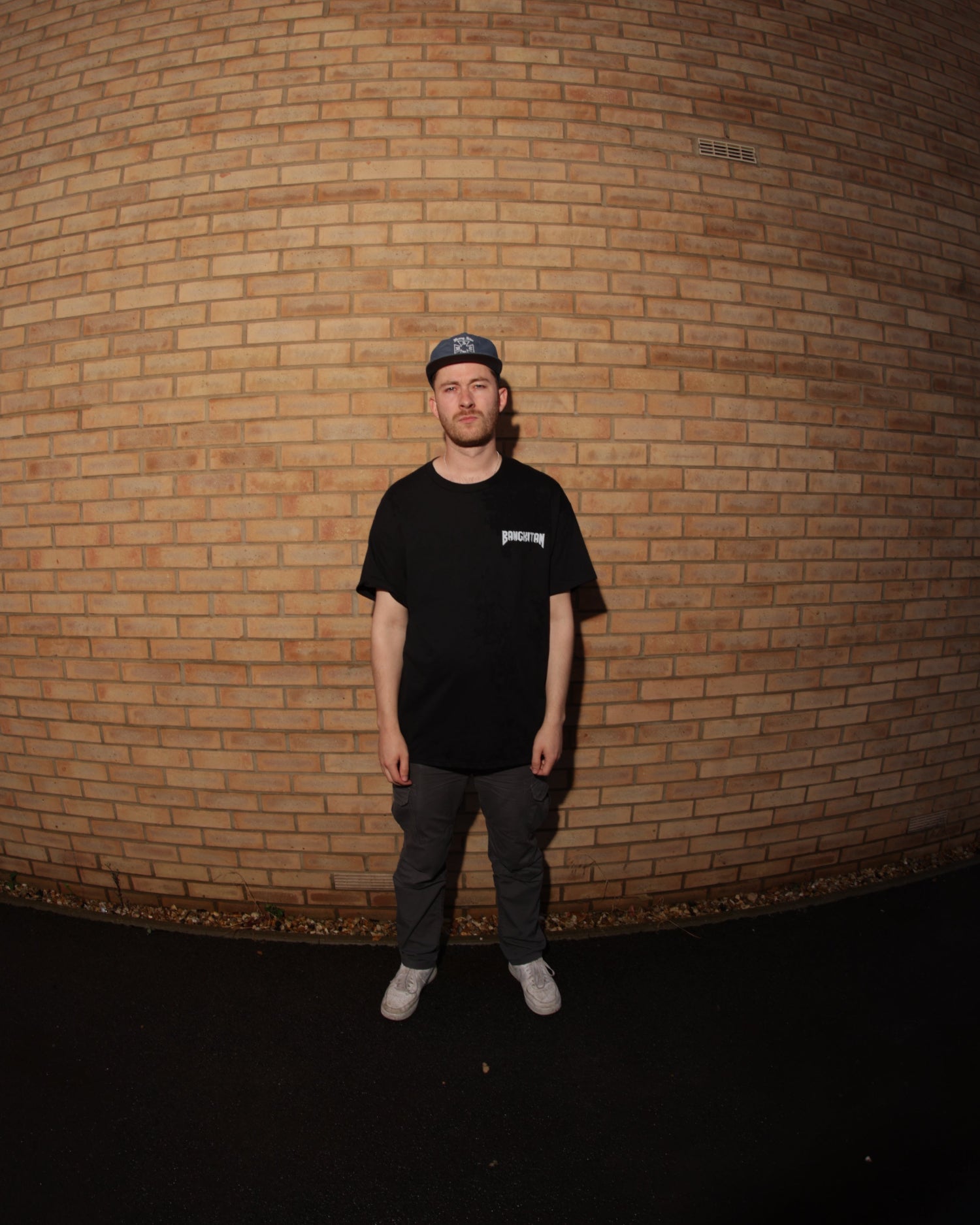 Man wearing a black BANGKITAN t-shirt and cap standing against a brick wall.