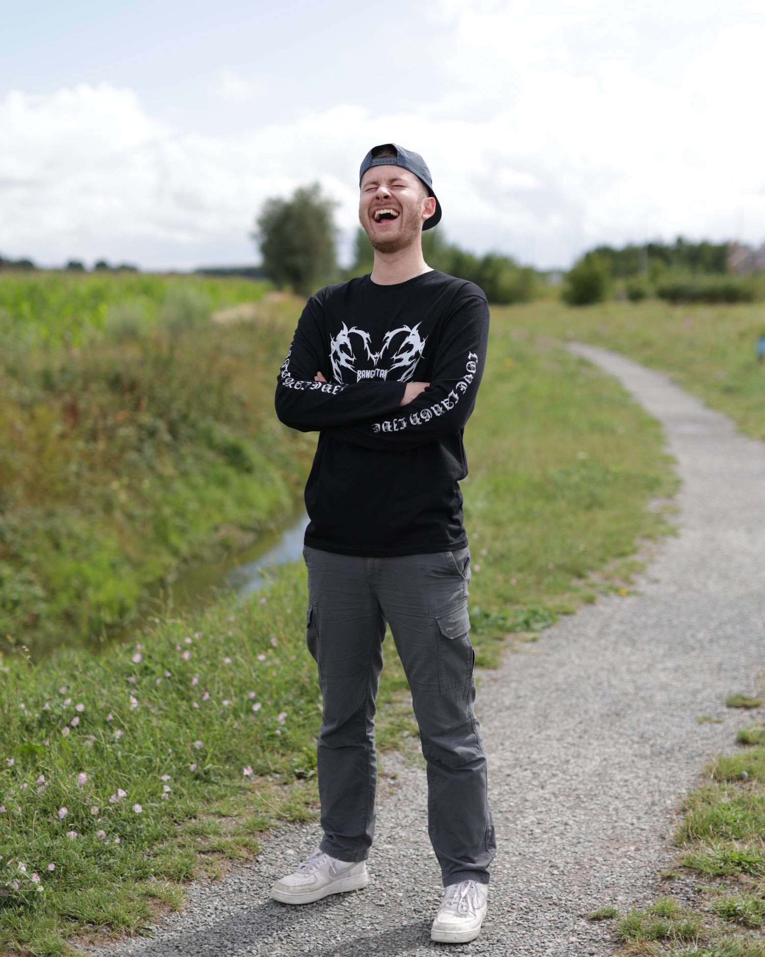 Person wearing a black long-sleeved BANGKITAN T-shirt standing on a gravel path with grass and trees in the background.